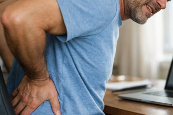 Muškarac sedi u kancelarijskoj stolici i drži se za donji deo leđa zbog bola izazvanog dugo sedenje tokom rada za računarom.
